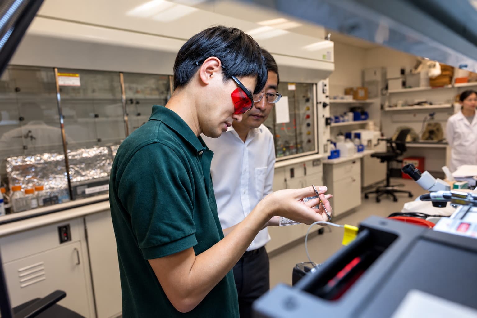 A researcher wearing red laser-safety goggles works at an optical rig while a colleague looks on